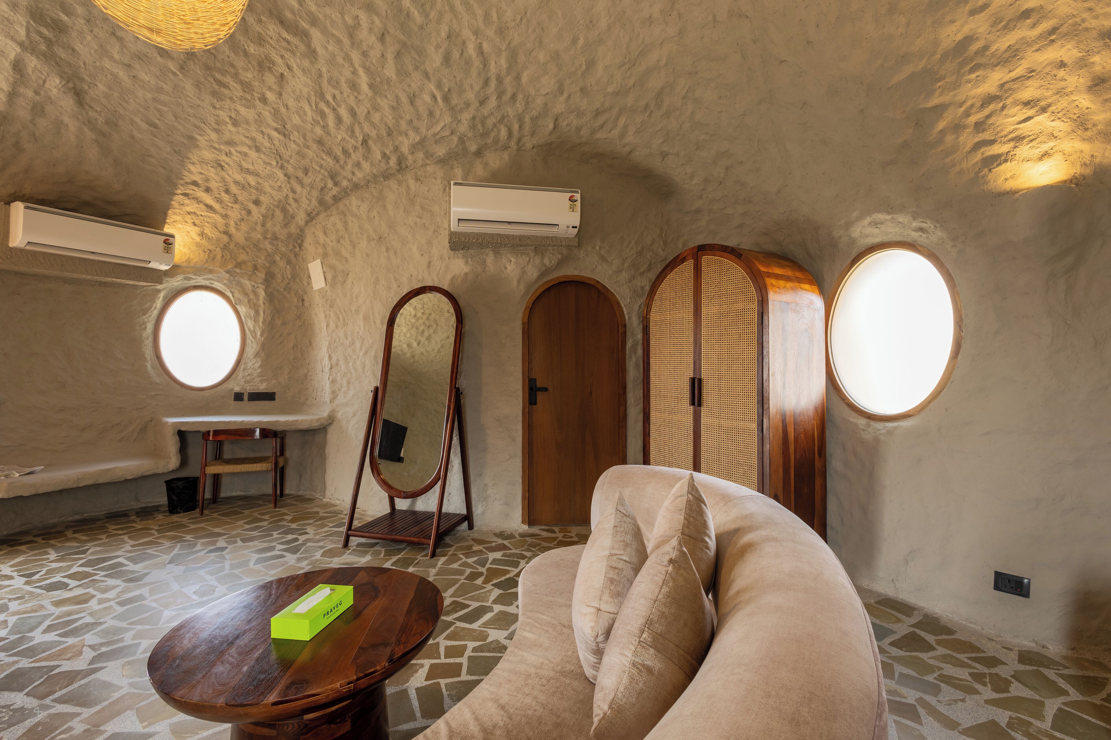 Une pièce chaleureuse avec un sol en pierre, une table ronde, un canapé beige, une porte en bois et une fenêtre ronde.