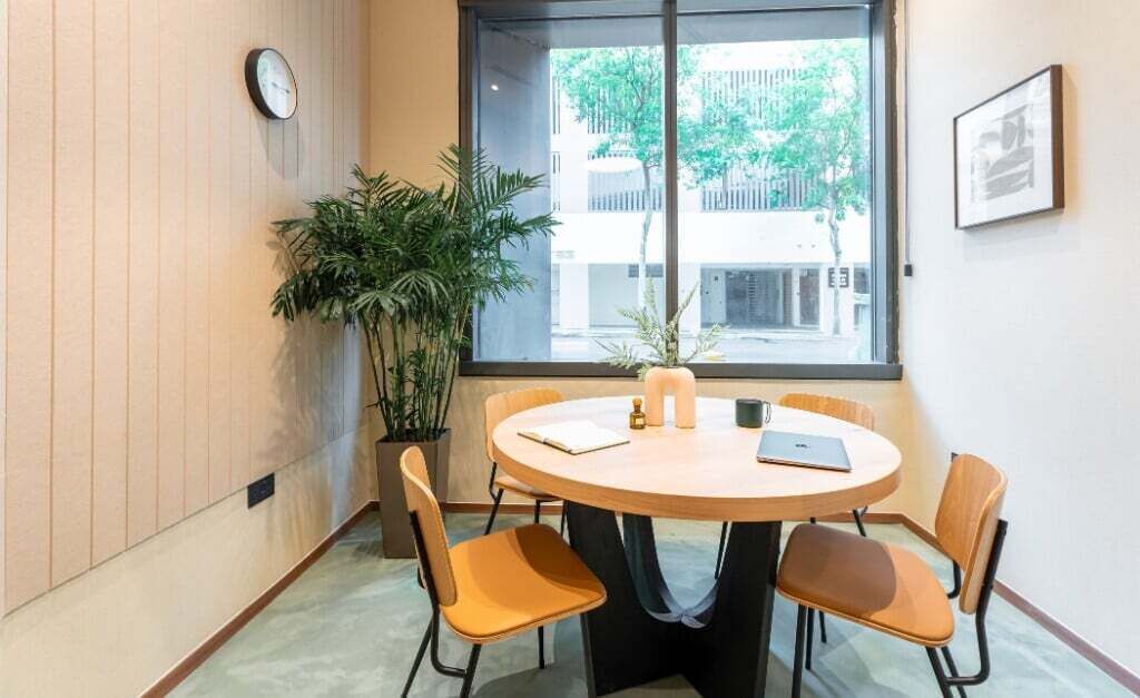 A conference room with a round table, four chairs, a clock, a potted plant, and a window with a view of buildings and trees.