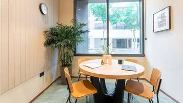 A conference room with a round table, four chairs, a clock, a potted plant, and a window with a view of buildings and trees.