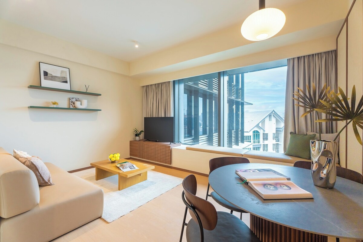 A modern living room with a sofa, coffee table, and dining table. There is a TV mounted on the wall, a window with curtains, and a plant on the windowsill.