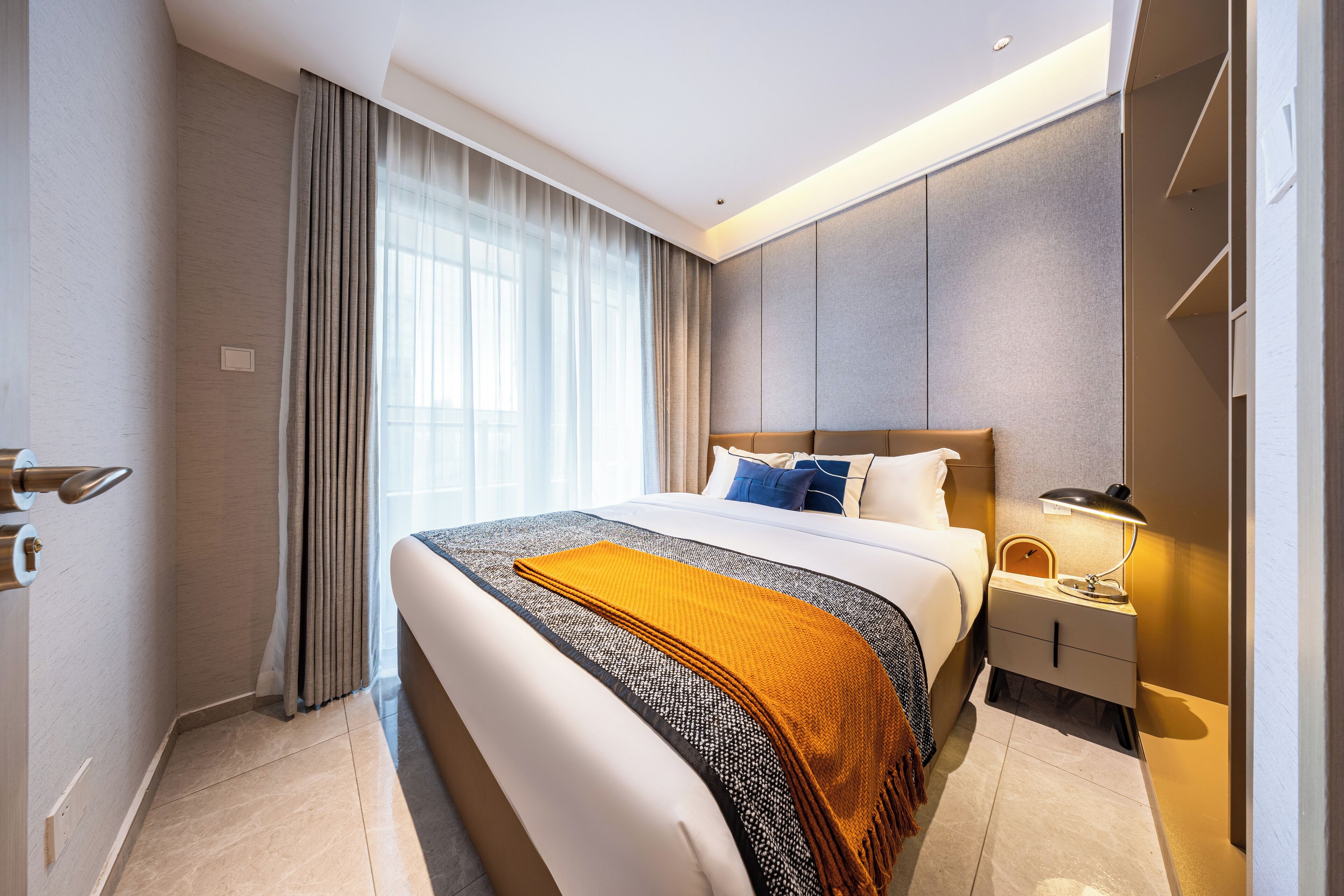 A modern hotel room with a large bed, a bedside table with a lamp, and a view of the city through the window.