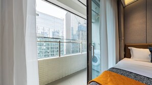 A hotel room with a bed, a window with a view of buildings, and a balcony with a railing. - Gangxiahui Hotel Apartments (Shenzhen)