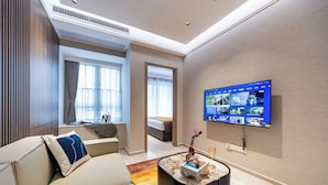 A modern hotel room with a large flat-screen TV mounted on the wall, a comfortable sofa with a decorative pillow, a small coffee table with a book and a vase, and a view of the adjacent bedroom through a doorway. - Gangxiahui Hotel Apartments (Shenzhen)