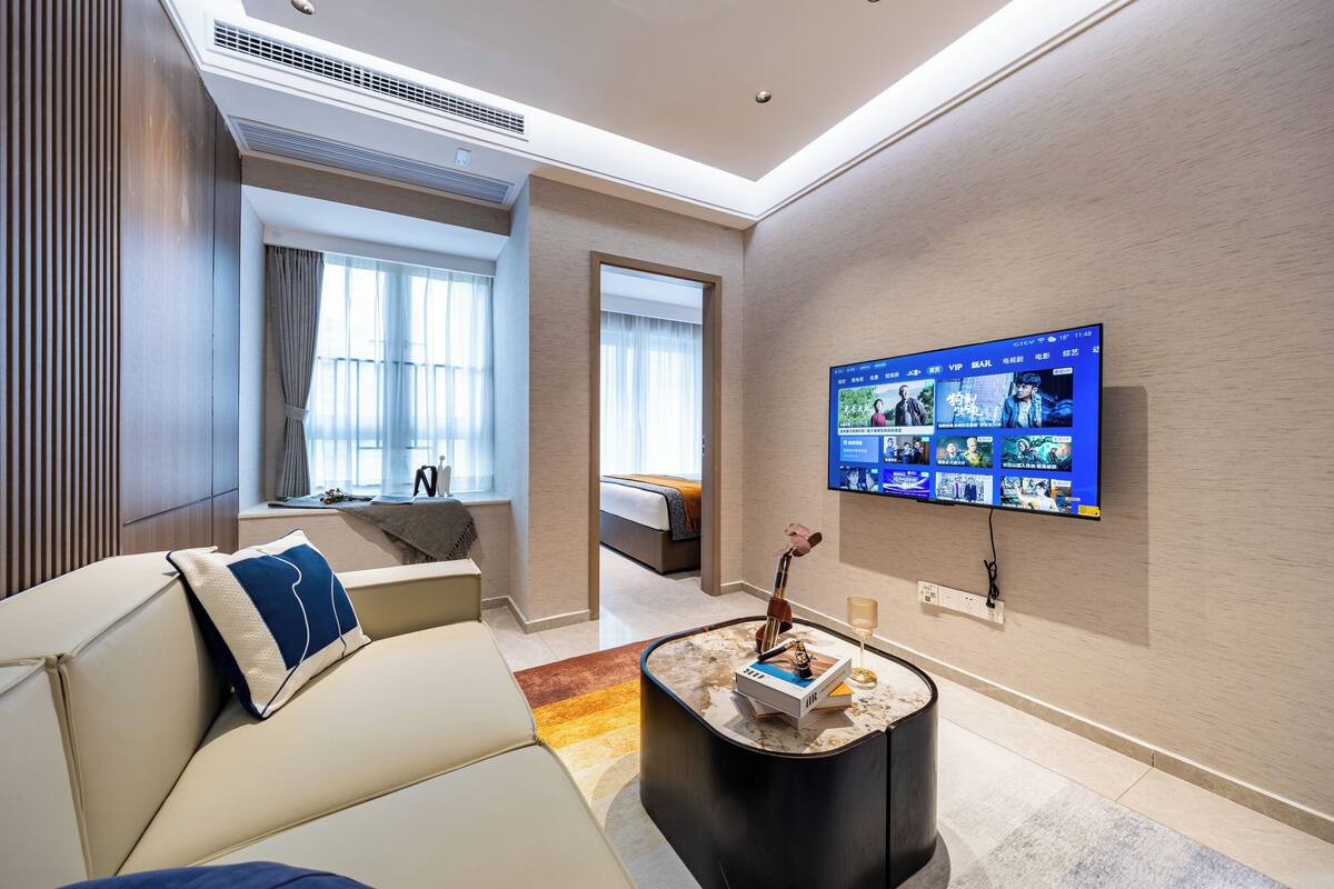 A modern hotel room with a large flat-screen TV mounted on the wall, a comfortable sofa with a decorative pillow, a small coffee table with a book and a vase, and a view of the adjacent bedroom through a doorway.