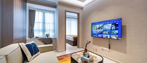 A modern hotel room with a large flat-screen TV mounted on the wall, a comfortable sofa with a decorative pillow, a small coffee table with a book and a vase, and a view of the adjacent bedroom through a doorway.