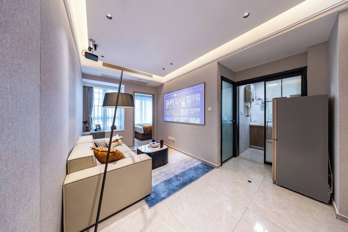 A modern hotel room with a sofa, a coffee table, a TV mounted on the wall, and a refrigerator.