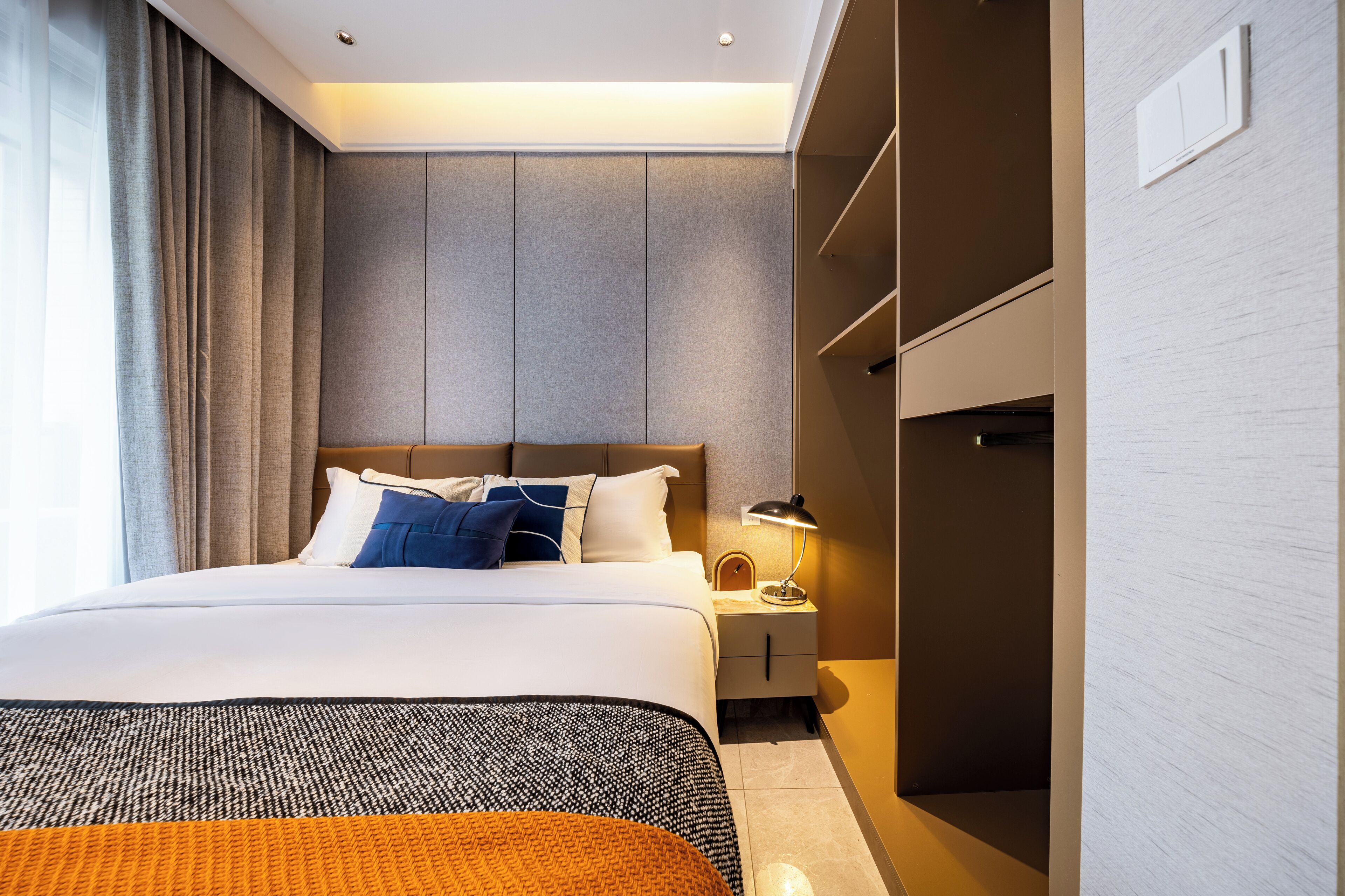 A modern hotel room with a large bed, a nightstand with a lamp, and built-in storage.
