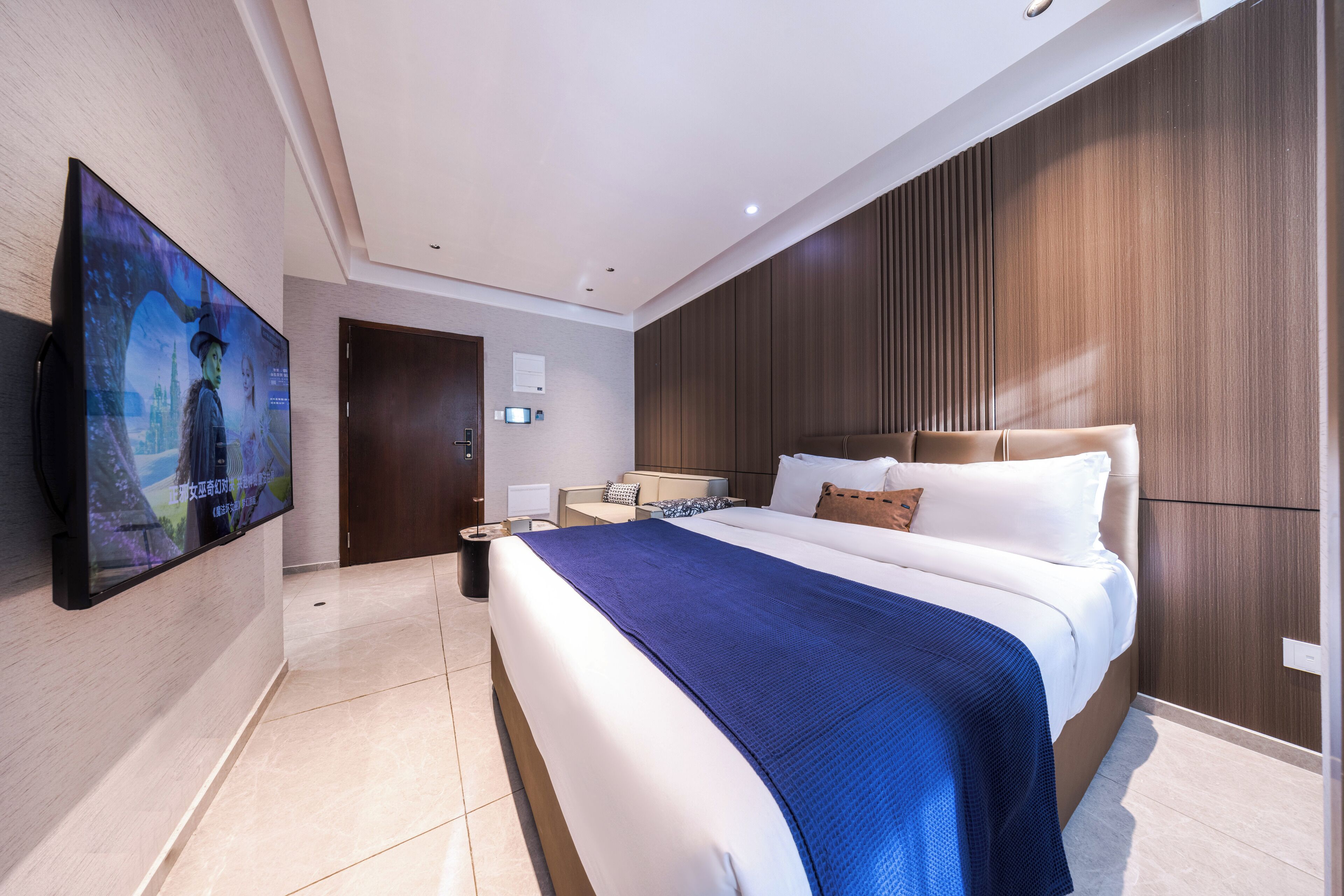 A modern hotel room with a large bed, a flat-screen TV mounted on the wall, a wooden panel wall, and a nightstand with a lamp.