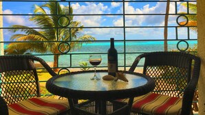 Une table ronde avec une bouteille et un verre, deux chaises en osier et une vue sur l’océan à travers une clôture en métal.