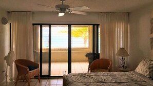 Une chambre d’hôtel avec un lit, deux chaises, un ventilateur de plafond, une lampe et une vue sur la plage, accessible par une porte coulissante.
