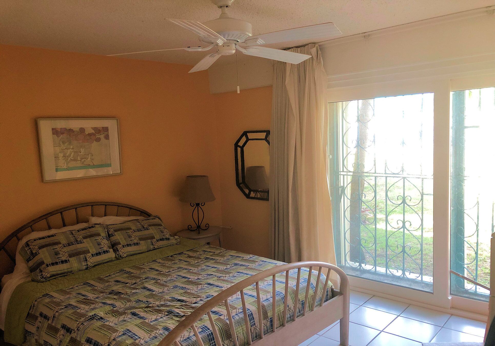A bedroom with a bed, a ceiling fan, a window with a view of greenery, a nightstand with a lamp, and a framed picture on the wall.