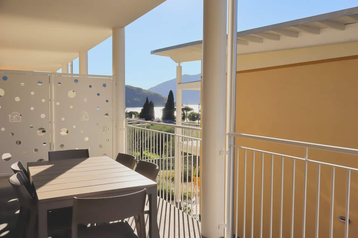 Uma varanda com mesa e cadeiras de madeira, corrimão branco e vista para montanhas e vegetação.