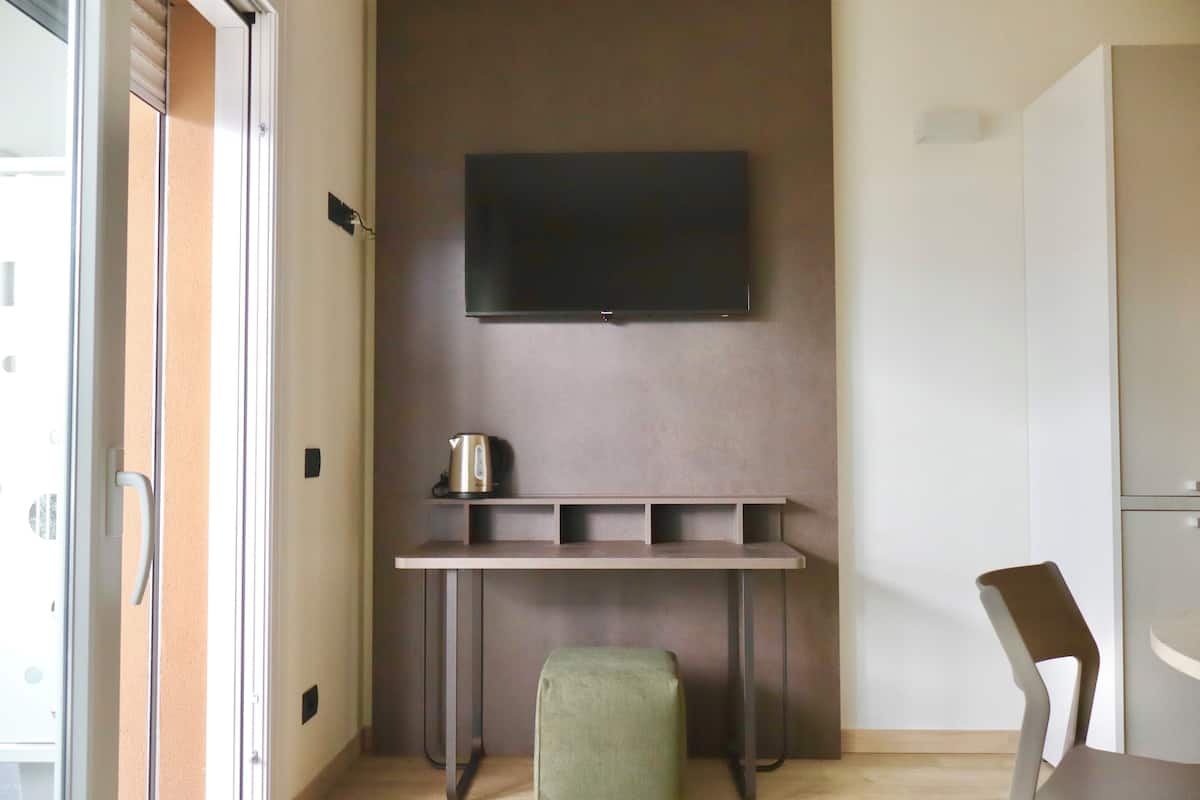 Quarto com televisão de tela plana na parede, uma mesinha com chaleira, um banquinho verde e uma cadeira.