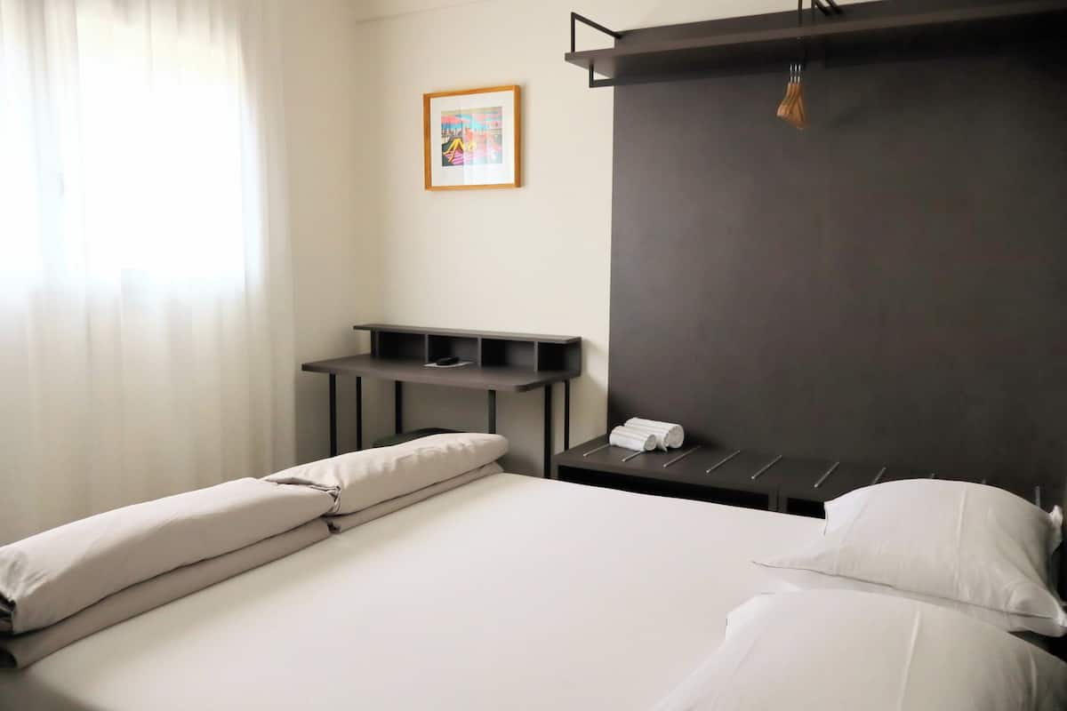 Quarto de hotel com cama, escrivaninha, cadeira, mesa pequena, luminária pendente e quadro na parede.
