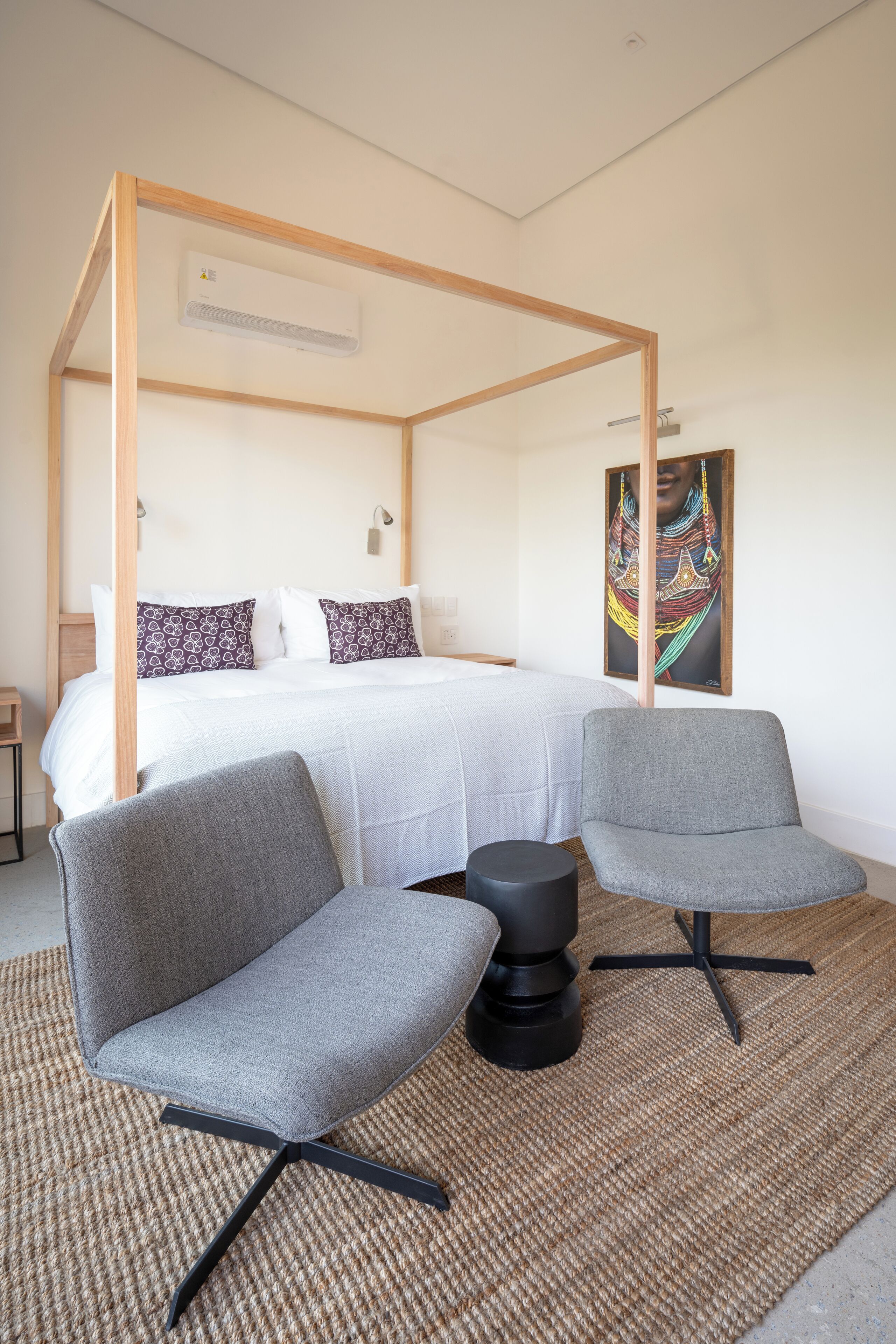 Un dormitorio moderno con una cama con dosel, dos sillones grises, un tapete tejido y una obra de arte colorida en la pared.