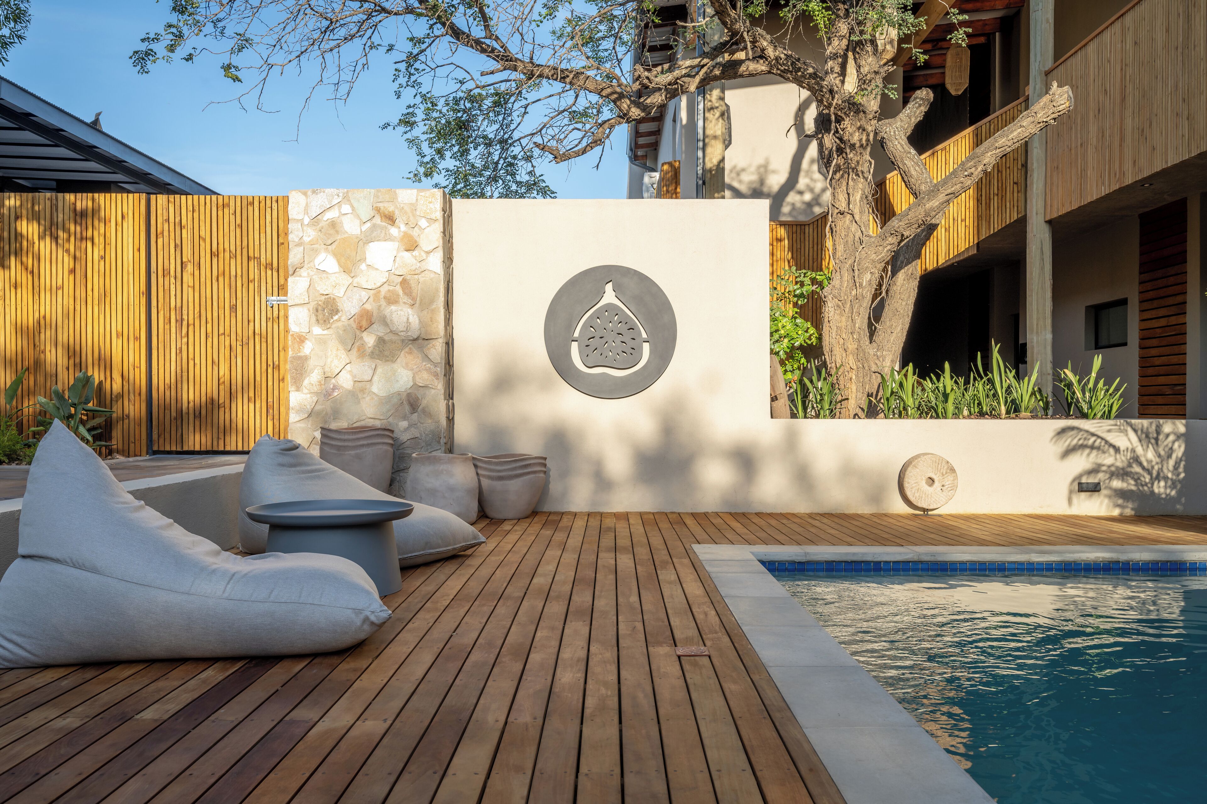 A wooden deck with a pool, stone wall, and a tree.