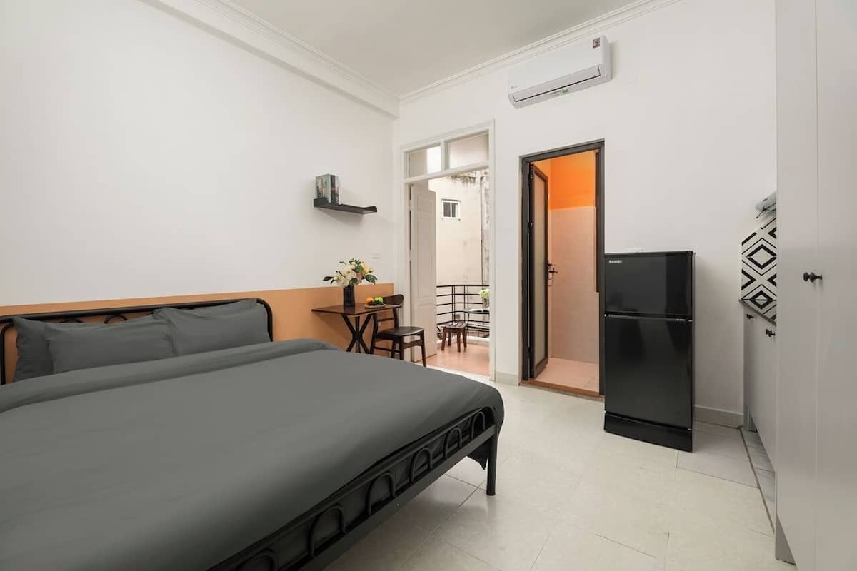 Een slaapkamer met een bed, een klein tafeltje met stoelen, een koelkast en een deur die leidt naar een balkon.