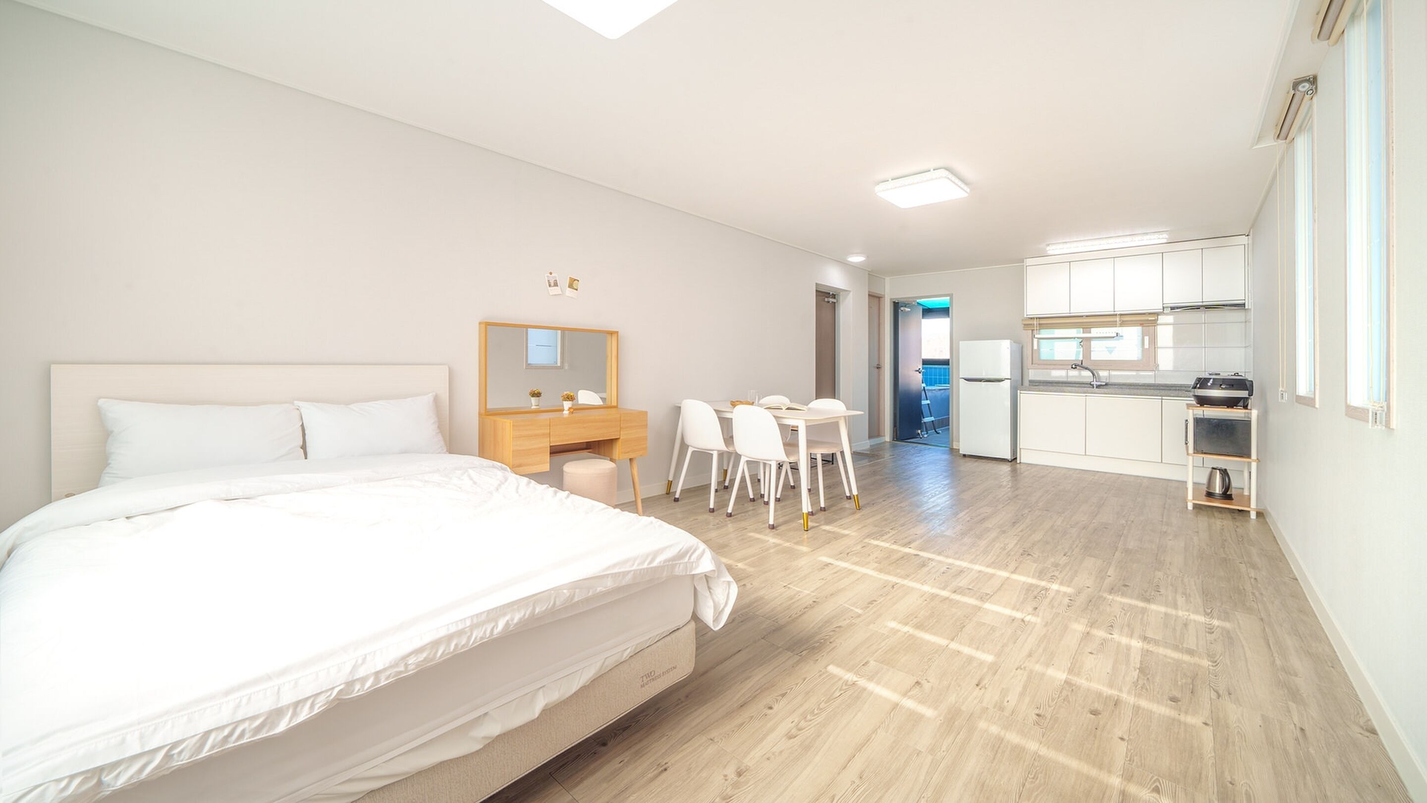 A modern hotel room with a large bed, a desk with chairs, a small kitchen area, and a bathroom visible through an open door.