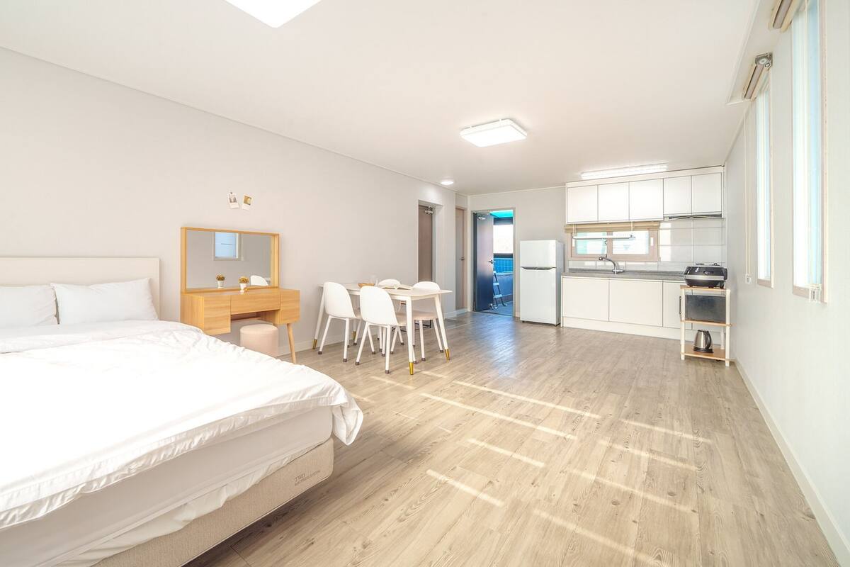 A modern hotel room with a large bed, a desk with chairs, a small kitchen area, and a bathroom visible through an open door.