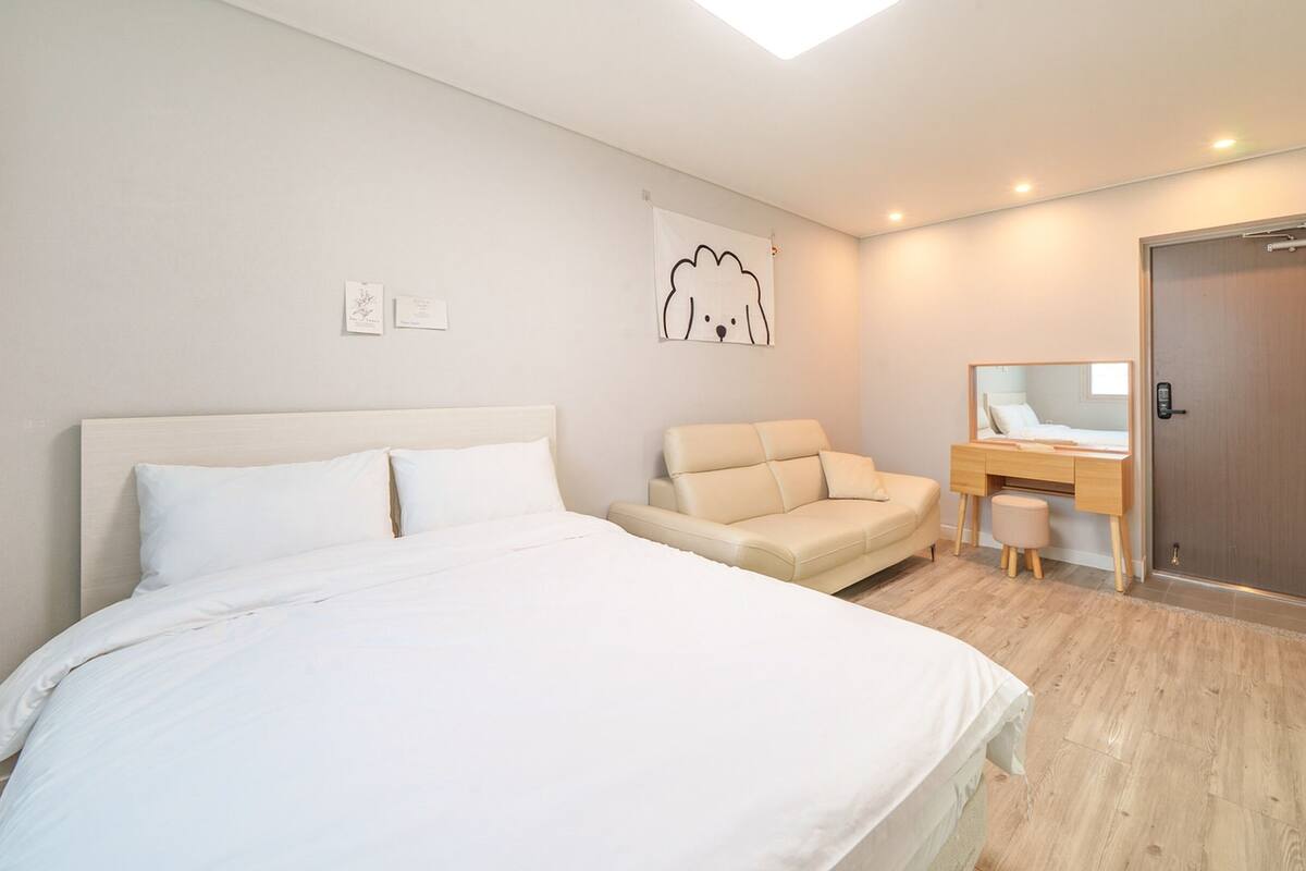 A modern bedroom with a large bed, a sofa, a desk with a mirror, and a door.