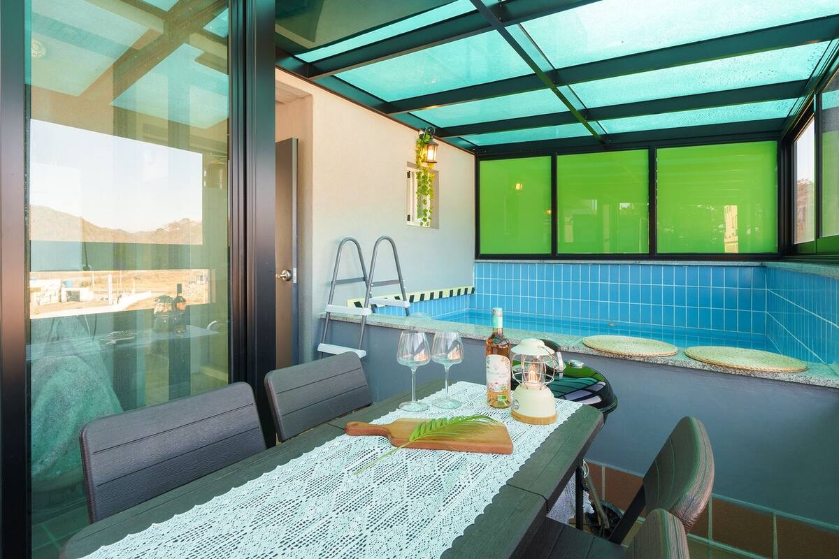 A dining area with a table set for two, a view of a swimming pool, and a glass-enclosed space with a view of the outside.