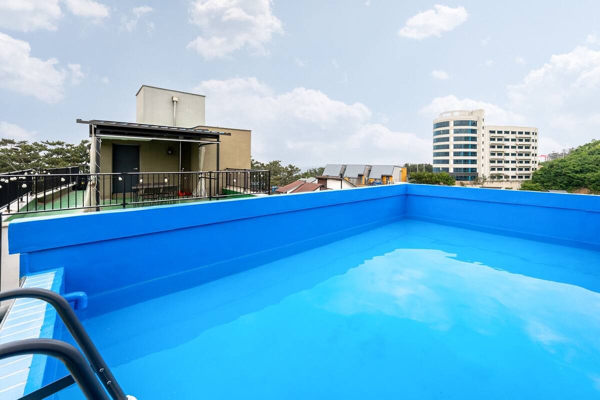 A rooftop swimming pool with a blue interior, surrounded by a black railing and a small outdoor seating area.