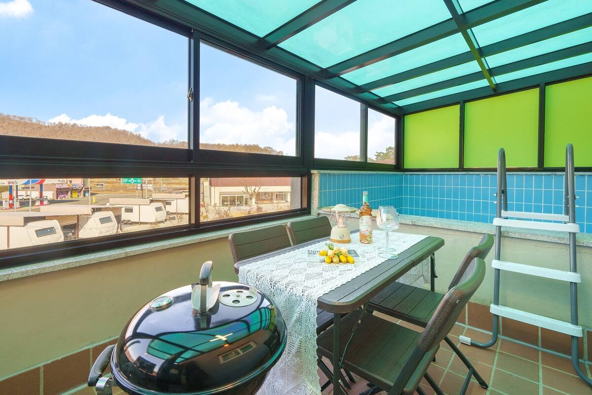 A balcony with a table, chairs, a grill, and a view of a parking lot and buildings.