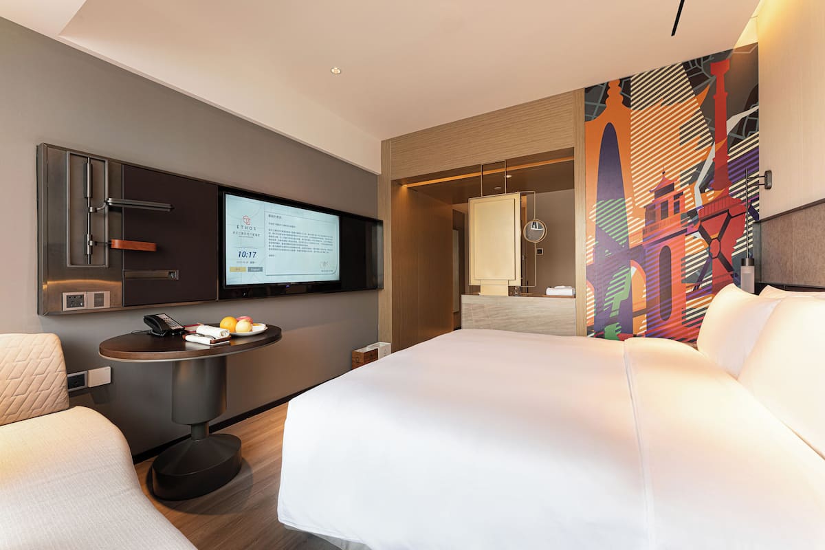 A modern hotel room with a large bed, a desk with a laptop, a television, and a colorful mural on the wall.