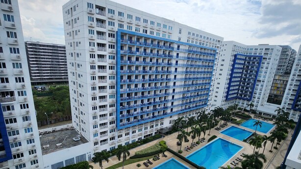 A high-rise hotel with multiple balconies, a central swimming pool, and a landscaped area with palm trees. - Spacious 2BR at Sea Residences Near MOA - Family Stay (Pasay)