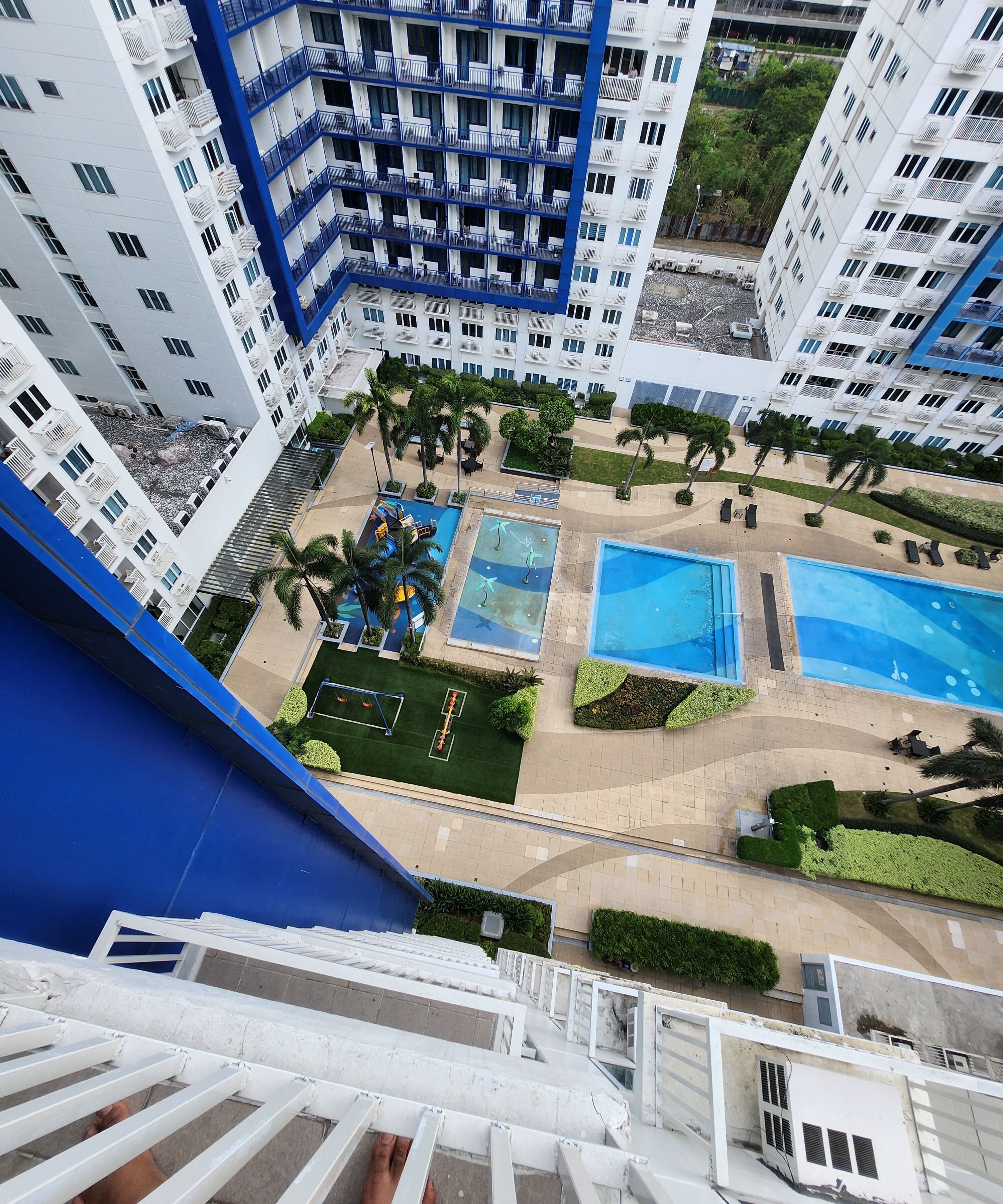 A high-rise building with multiple balconies, a swimming pool area with sun loungers, and a well-maintained garden with palm trees.