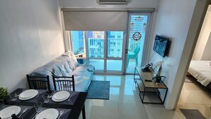 A modern living room with a sofa, dining table with chairs, a TV, and a balcony with a view of buildings. - Spacious 2BR at Sea Residences Near MOA - Family Stay (Pasay)