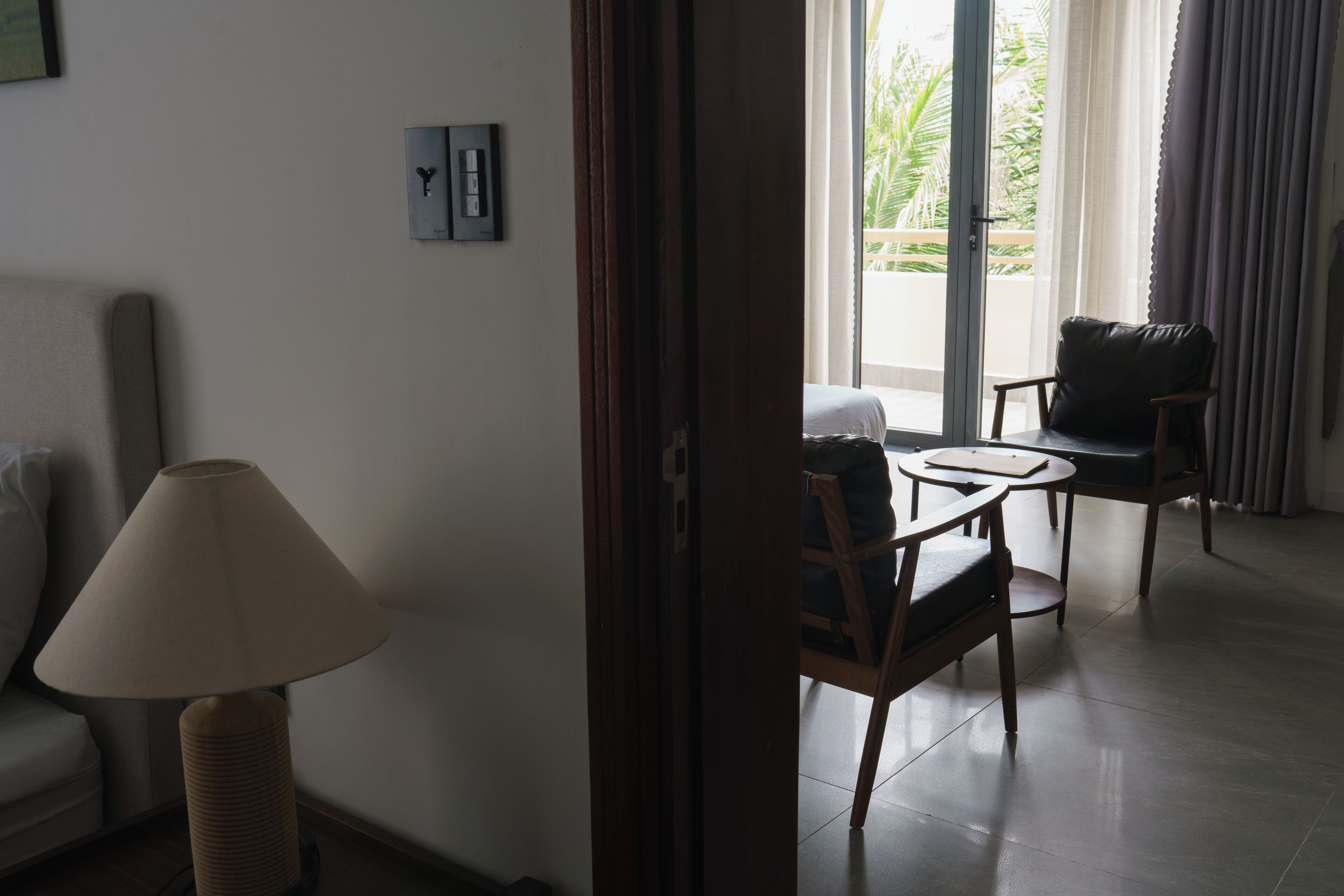 A hotel room with a sofa, a table, two chairs, a lamp, and a view of the outdoors.