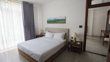 A neatly made bed in a hotel room with a painting on the wall, a nightstand with a lamp, and a view of the room through an open door.