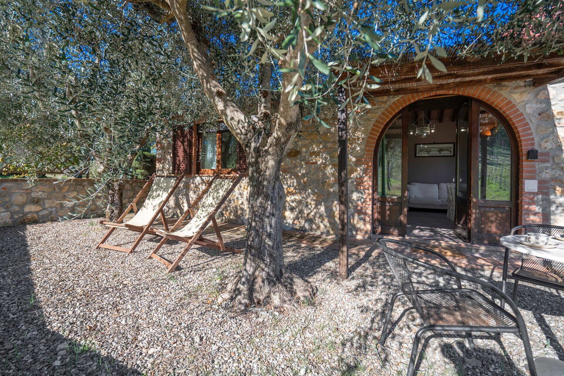 Un espace extérieur aménagé dans un style rustique, avec des chaises longues en bois et une table en métal, entouré d’oliviers et d’un mur en pierre.