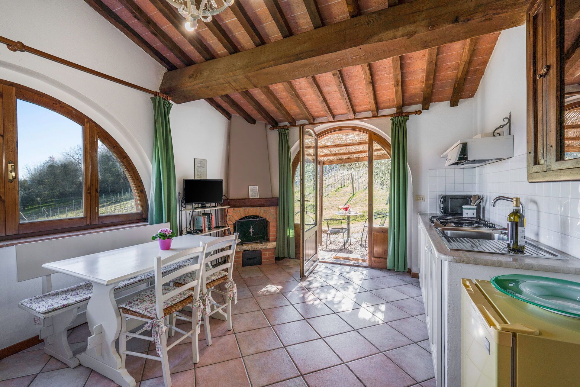 A rustic kitchen with a wooden ceiling, a dining area with a table and chairs, a TV, and a view of the outdoors through a window.