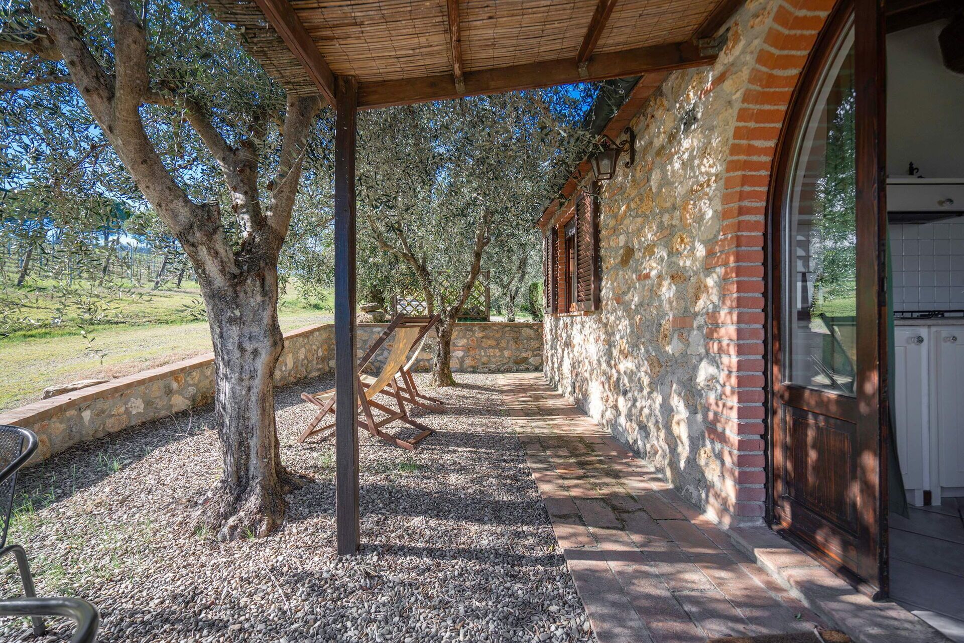 A covered outdoor area with stone walls and a wooden pergola, leading to a building with a stone facade and a window.