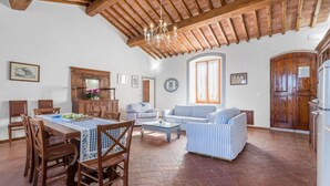 Ampla sala de estar com mesa de jantar, cadeiras, sofás e cozinha com armários de madeira e geladeira.