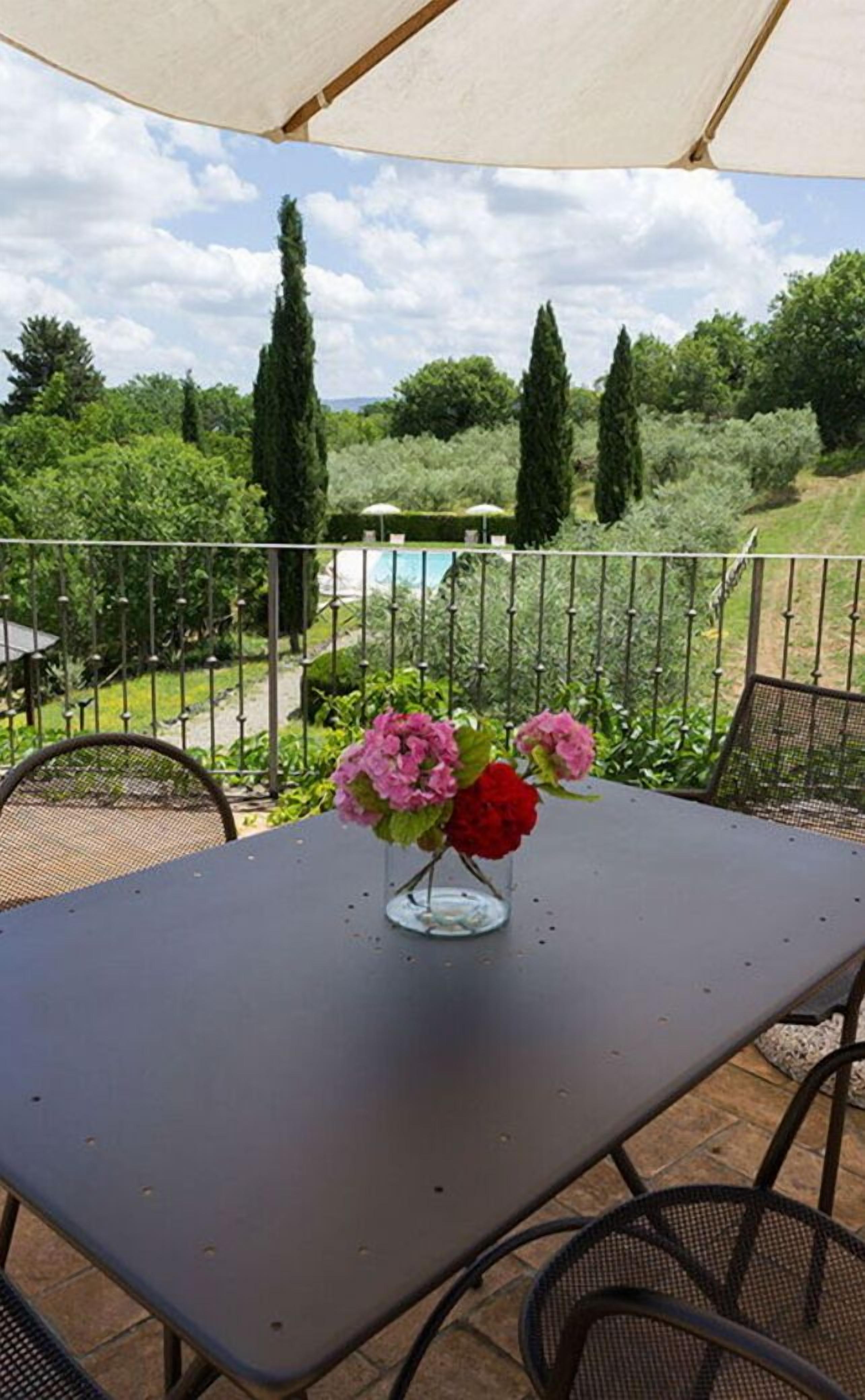 Een terras buiten met een tafel en stoelen, een vaas met bloemen, en uitzicht op een zwembad en groen.
