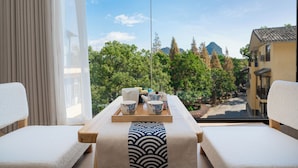 A room with a view of a mountain and trees, a wooden table with a tray of tea set, and two white cushioned chairs.