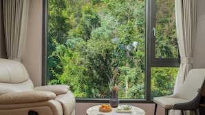 A room with a large window, a round table with a vase and fruit, and two chairs.