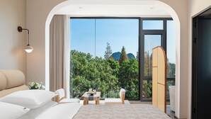 A modern hotel room with a large sliding glass door leading to a balcony, a bed, a small table with a vase of flowers, and a seating area with chairs.