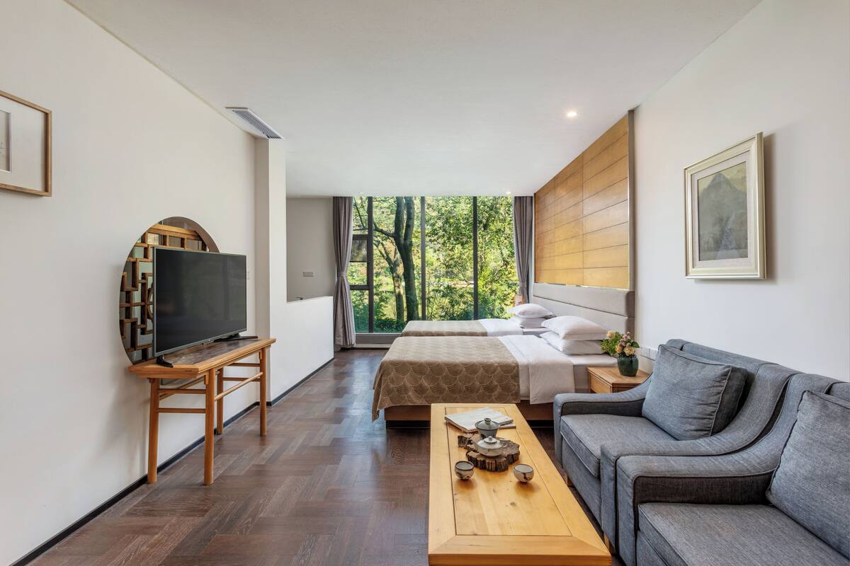 Ein modernes Hotelzimmer mit einem großen Bett, einer Couch, einem Fernseher auf einem Holzboden und Blick auf Bäume durch das Fenster.