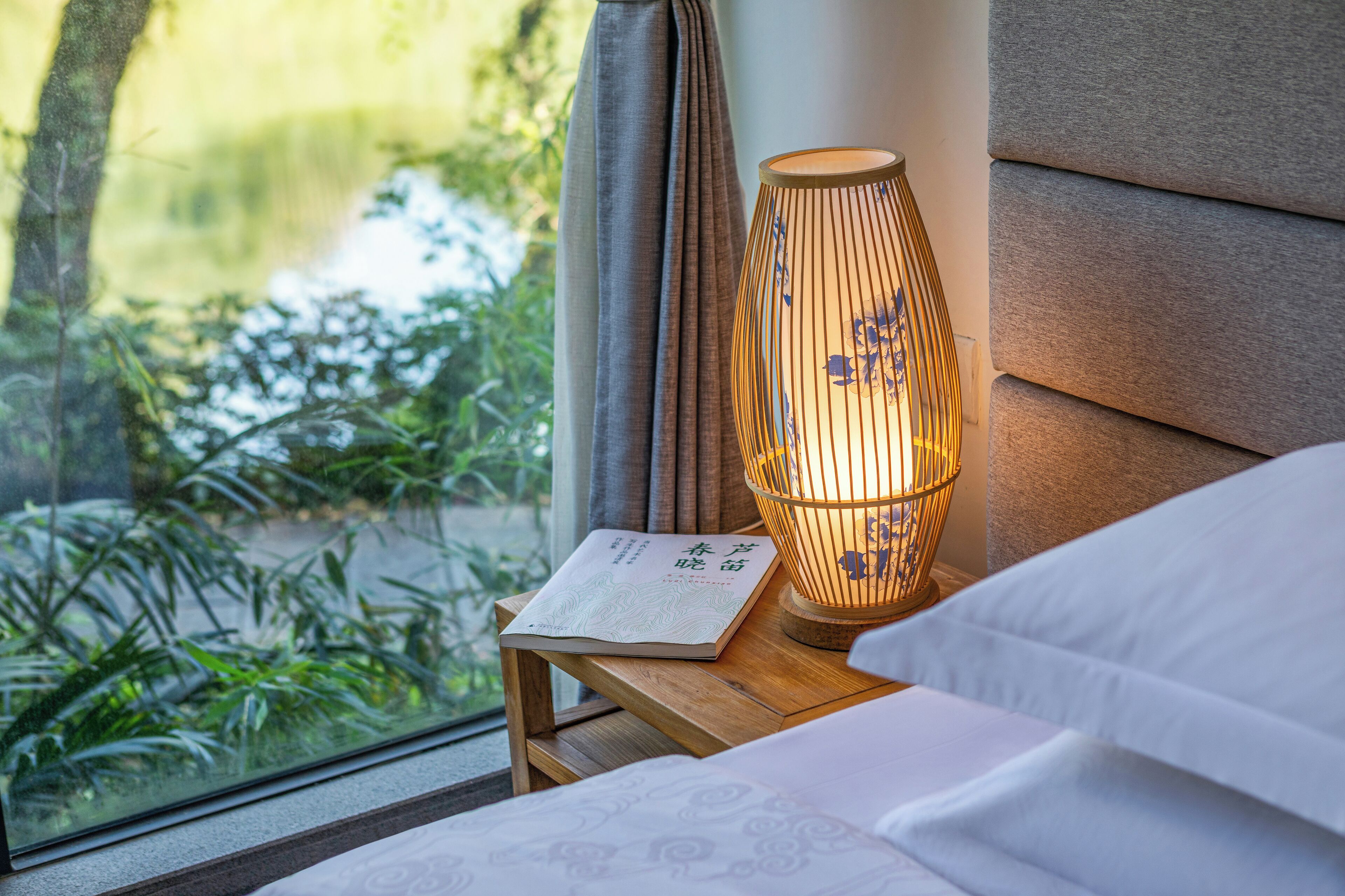 Una cama bien hecha con edredón blanco, una mesita de noche con un libro y una lámpara, y vistas a zonas verdes por una ventana.