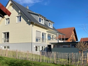 A two-story residential building with a sloped roof, a visible front porch, and a white picket fence.