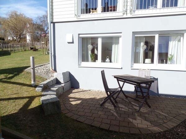 A two-story building with a patio area featuring a table and chairs, a small garden, and a white exterior.