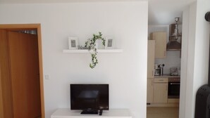 A living room with a TV on a white stand, a small shelf with decorative items, and a kitchen in the background.