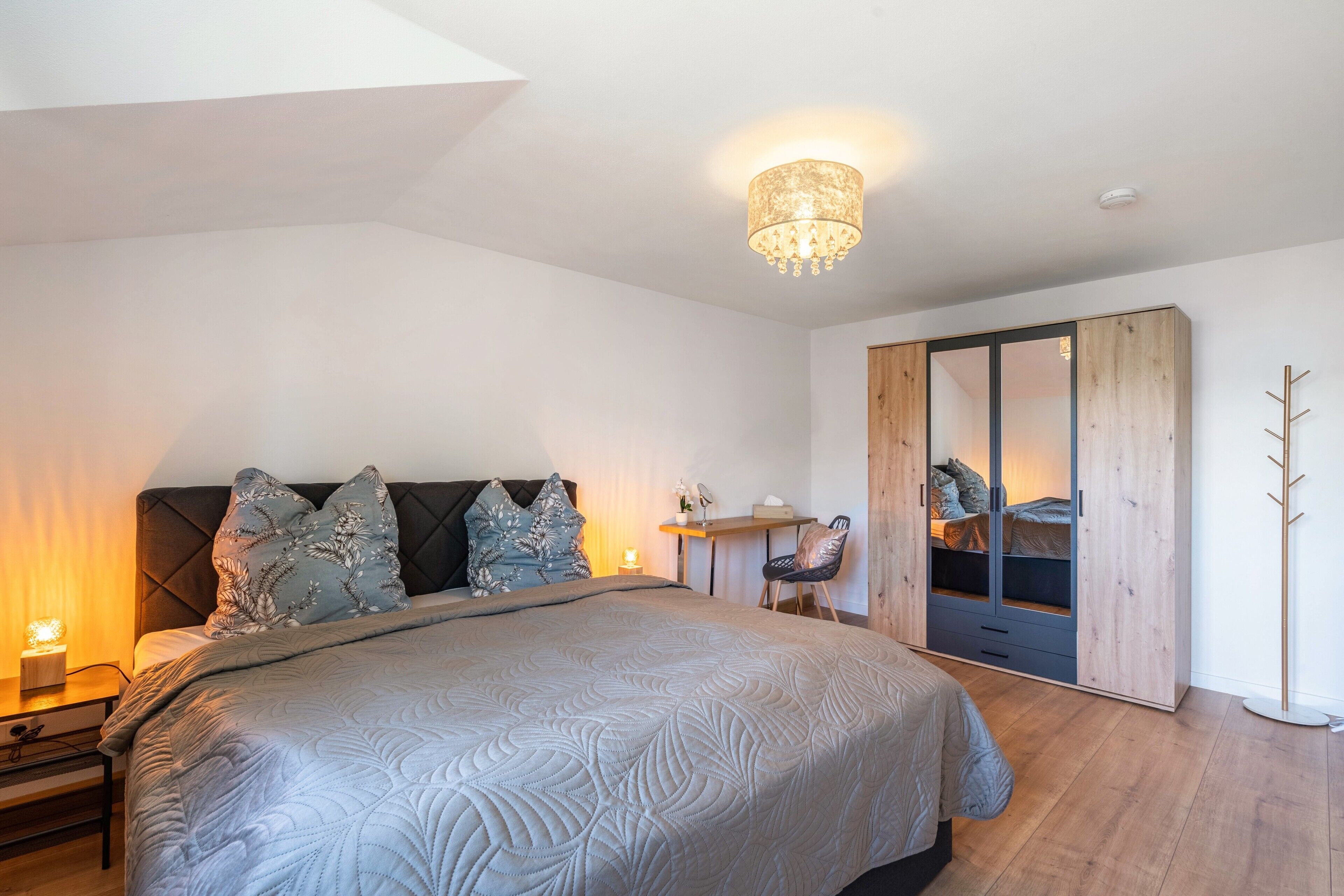A bedroom with a large bed, a nightstand, a desk, a chair, a wardrobe, and a coat rack.