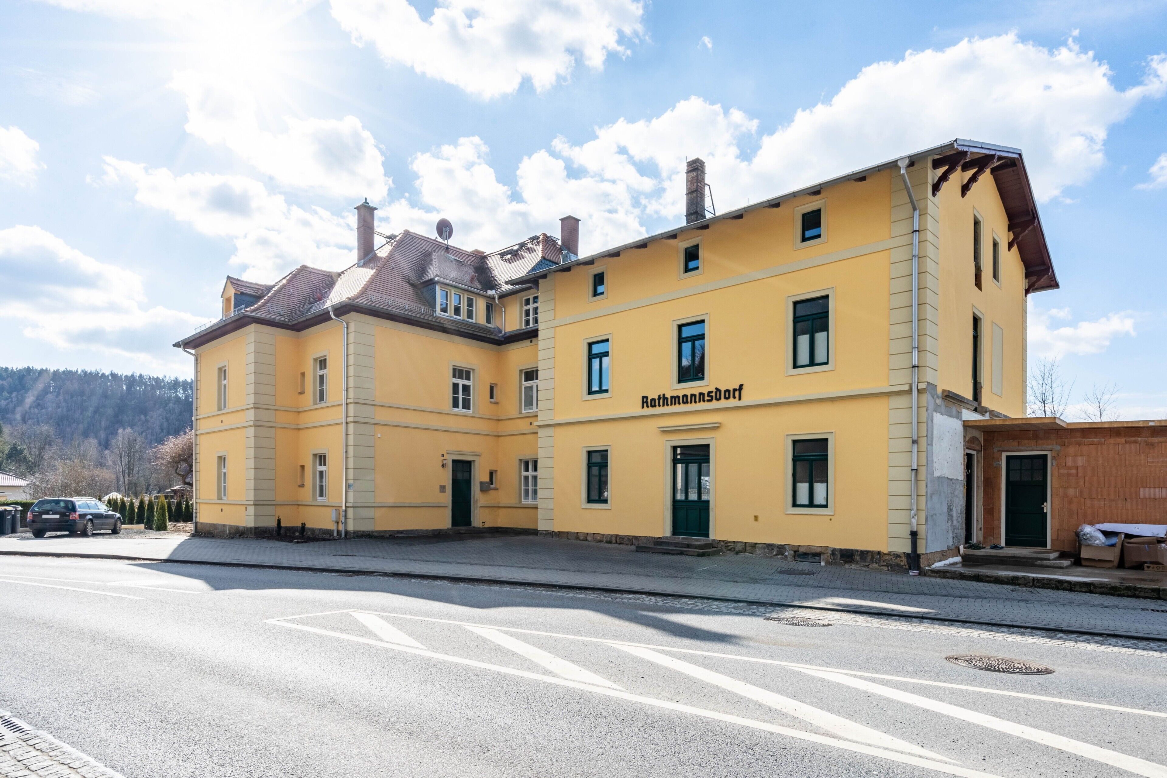Ein gelbes Gebäude mit der Aufschrift 'Rathmannsdorf' an der Fassade, eine gepflasterte Straße davor und ein klarer Himmel.