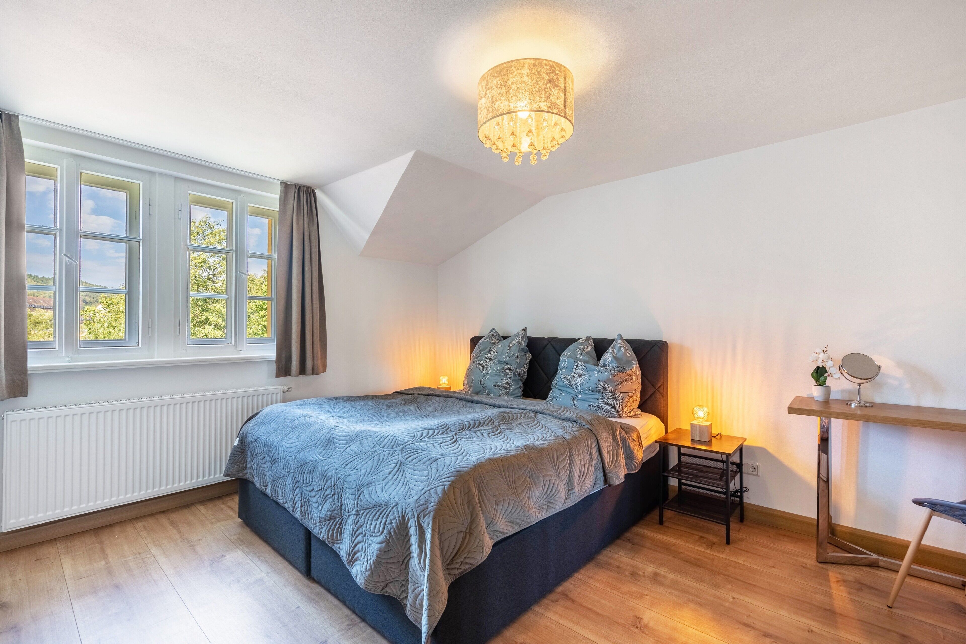 A bedroom with a bed, a nightstand, a mirror, a window with curtains, and a ceiling light fixture.