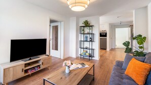 Un salon moderne doté d’un téléviseur à écran plat, d’un meuble de rangement en bois et d’une table basse sur laquelle reposent un magazine et un bouquet de fleurs.